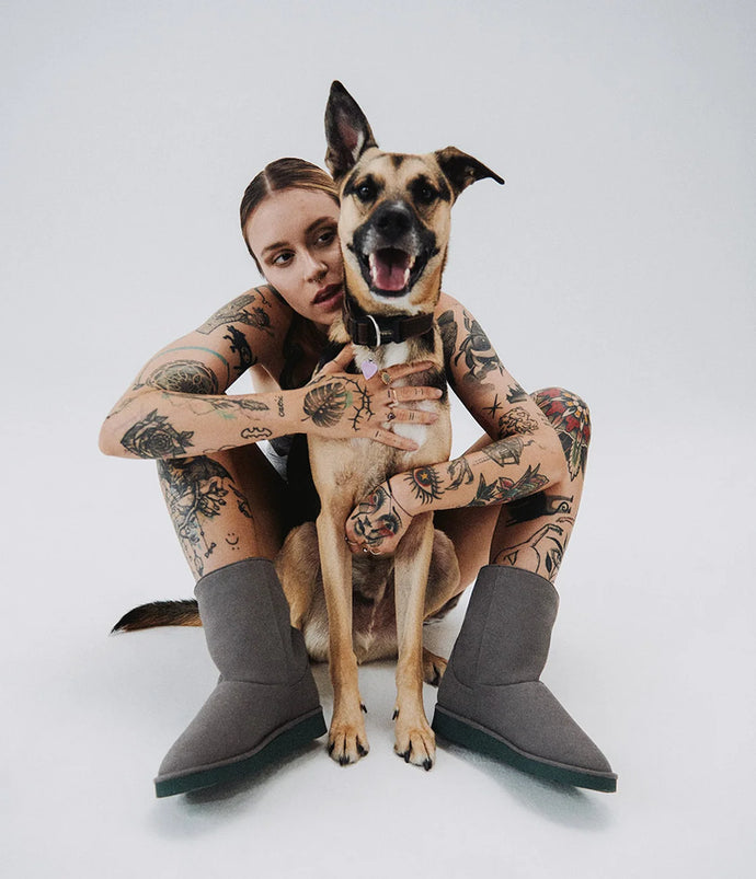 Woman with tattoos sitting on the floor with a dog, wearing gray boots.