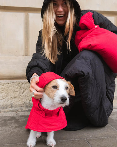 Woman in black coat with black skirt petting a dog wearing a red coat on a stone pavement.