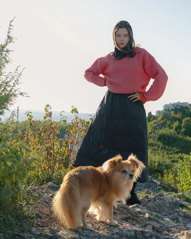 Woman in a pink top and black skirt standing with a dog on a rocky path with greenery in the background