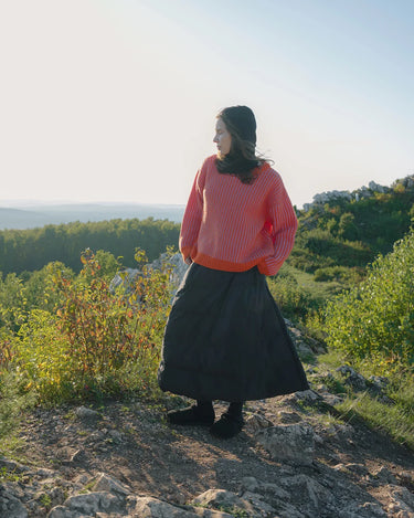 Person wearing a pink sweater and black skirt standing on a rocky outcrop with greenery and a clear sky in the background.