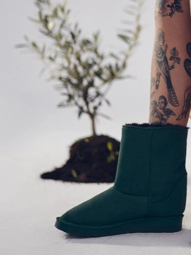 Person wearing dark green slip-on boots with a tattooed arm and legs on a light background