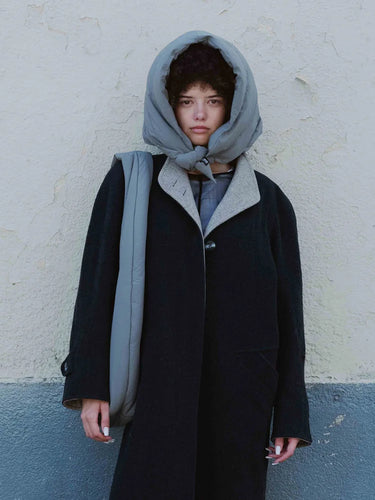 Person wearing a black coat with a hood against a plain wall.