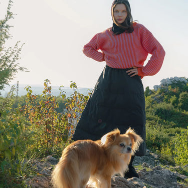 Woman in a pink top and black skirt standing with a dog on a rocky path with greenery in the background