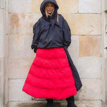 Person wearing a black hooded jacket and red puffer skirt against a stone wall.