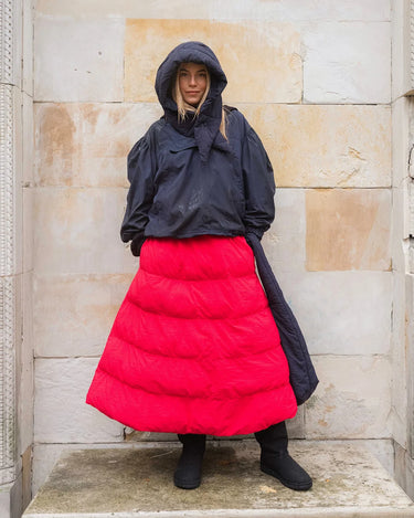 Person wearing a black hooded jacket and red puffer skirt against a stone wall.
